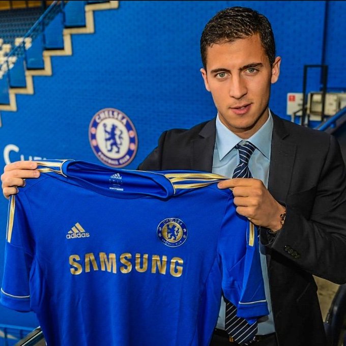 Eden Hazard holding up a blue Chelsea FC jersey featuring the Samsung and Adidas logos, posing in front of a blue stadium backdrop with the Chelsea FC crest.