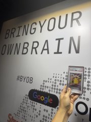A promotional display featuring the slogan 'BRING YOUR OWN BRAIN' with the hashtag #BYOB, alongside a Google device and a phone showing an image, with a hand reaching toward it.