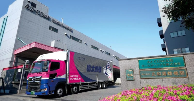 Photograph of an AZ.COM delivery truck parked outside the AZ-COM Centrepiece building in Japan, featuring pink and blue branding with Japanese text on the signage.