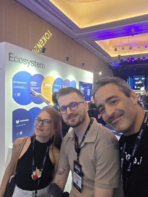 Three attendees at a cryptocurrency conference pose for a photo near a "TOKEN2049" branded booth featuring an "Ecosystem" display with logos for BNB Finance, USDM, and minswap, capturing a friendly meet-up moment at the event.