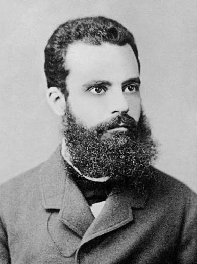 A black-and-white historical portrait of a bearded man in a dark suit and bow tie, likely from the 19th century.