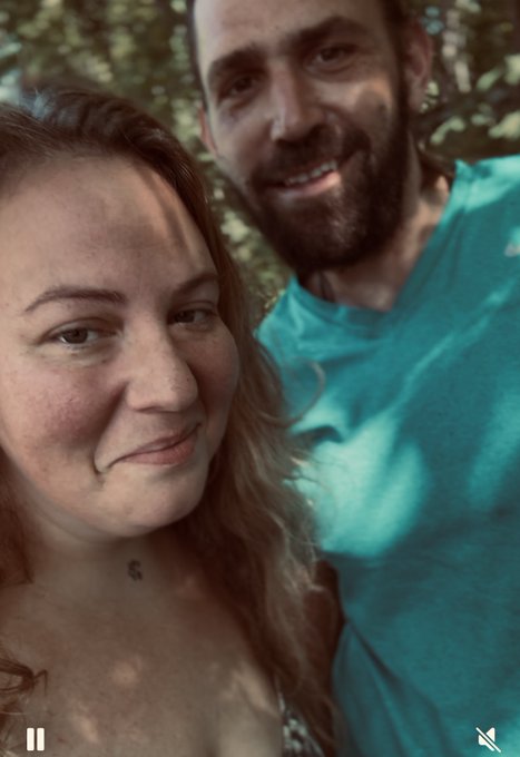 A selfie of a smiling couple outdoors among trees, with a woman in the foreground and a bearded man in a teal shirt behind her.