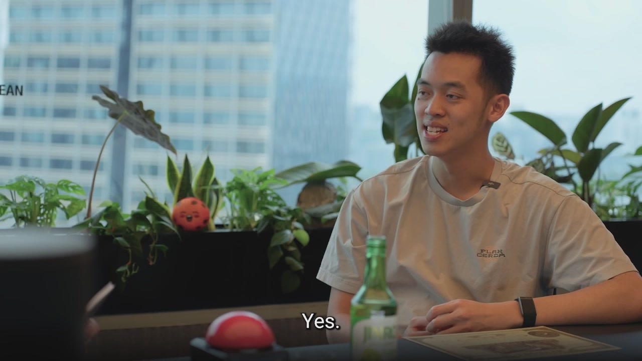 The image shows a man sitting at a table, smiling as he looks directly at the camera. The background features a window overlooking a cityscape, adding an urban touch to the scene. On the table, there are two potted plants - one with a red ball on top and another with a green plant. A bottle of beer is also present on the table. The image includes text that reads "it' s been the 2nd solana ecosystem call by @solBrothersPod since we announced we're coming to @solana with @ikadotxyz and @encrypt_xyz".