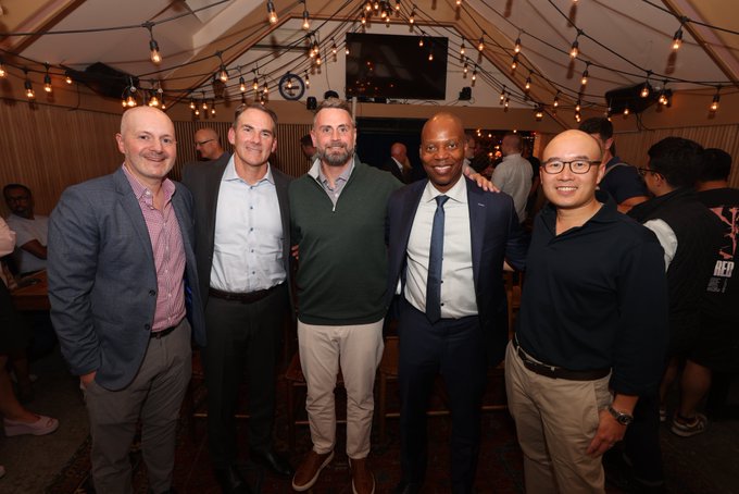 Five men in business casual attire posing together for a group photo at an indoor event venue decorated with string lights.