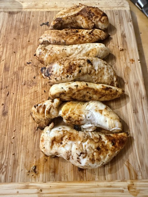 Seven seasoned and grilled chicken breasts arranged vertically on a wooden cutting board.