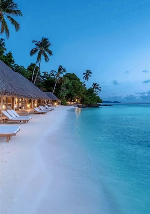A serene tropical beach scene at dusk with white sand, lounge chairs, thatched-roof huts, palm trees, and calm turquoise water under a deep blue sky.