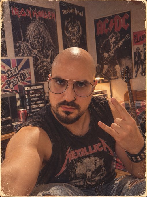 A bald man with a beard, glasses, and a nose ring wearing a Metallica t-shirt, making a rock hand gesture while taking a selfie in a room decorated with Iron Maiden, Motörhead, AC/DC, and Sex Pistols posters.