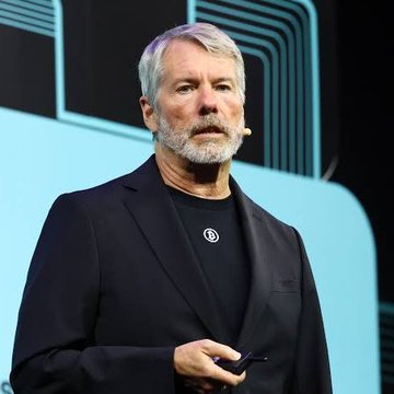 A man with grey hair and beard wearing a black blazer and dark t-shirt speaks on a stage with a teal background, holding a clicker or small device in his hand.