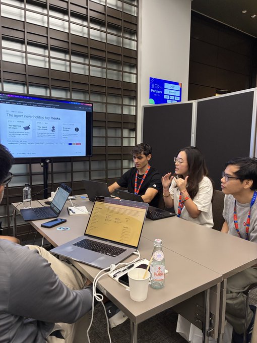 A group of hackers collaborates around laptops at an ETHGlobal Tokyo event, with a monitor displaying a slide titled "The agent never holds a key. It talks," reflecting the hackathon's focus on AI agents, security, and decentralized solutions.