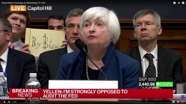 A screenshot from a Bloomberg TV live broadcast on Capitol Hill shows Janet Yellen, identified by the chyron "YELLEN: I'M STRONGLY OPPOSED TO AUDIT THE FED," testifying with a man in the background holding a "Buy Bitcoin" sign—the same photobomb famously captured in the original moment shared 9 years ago today. The S&P 500 ticker shows 2,440.98.