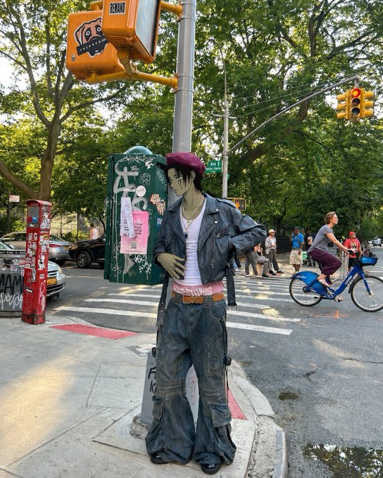 A street performer dressed in a purple cap, pink shirt, and denim outfit stands on a New York City street corner near a crosswalk, with a cyclist and pedestrians in the background.