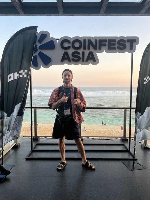 A man gives two thumbs up while posing in front of a large "COINFEST ASIA" sign at a beachside venue in Bali, with the ocean visible in the background. The caption captures his enthusiasm for the crypto conference experience at a beach club in Uluwatu, matching the caption's note about enjoying the event and interacting with the friendly organizers.