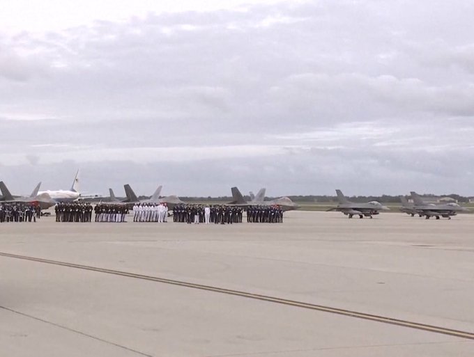Military personnel in formation stand on an airfield tarmac lined with fighter jets and other military aircraft under an overcast sky.