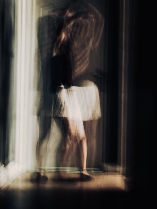 A blurred, ghostlike figure in a white skirt stands barefoot in a softly lit doorway, hair tossed in motion, embodying the fleeting passage between presence and absence that the artist describes. The painterly motion and dissolving form visually echo the post's meditation on fluidity, change, and the space between two silences.