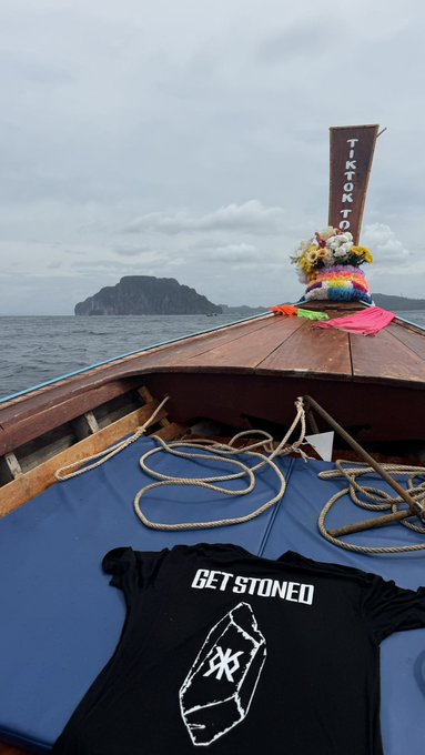 The bow of a wooden boat on the water holds a black t-shirt reading 'GET STONED' with a coffin graphic, while colorful floral offerings are tied to the boat's bow against a backdrop of a rocky island.