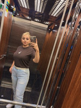 A woman takes a mirror selfie in a wood-paneled elevator, wearing a black Nike t-shirt and light blue jeans, holding a phone with a patterned case. The polished, reflective surfaces of the elevator echo the post's theme of mirroring and self-reflection, though she appears to be sharing a moment rather than displaying the artwork or cryptocurrency referenced in the caption.