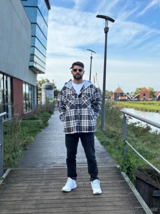 A man wearing sunglasses, a plaid shirt, black pants, and white sneakers standing on a wooden boardwalk.