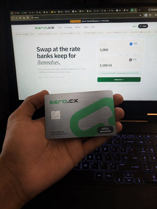 A hand holding a metallic 'sera.cx' debit card powered by XSGD Singapore, displayed in front of a laptop showing the sera.cx website with text 'Swap at the rate banks keep for themselves.'