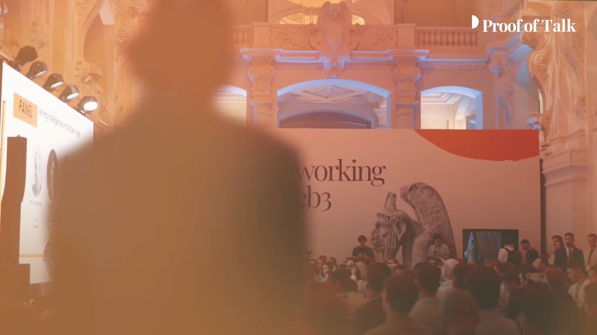 The image shows a large crowd gathered for an event at the Louvre museum. The crowd consists of people standing around a stage where a speaker is giving a presentation. The backdrop features a large banner with the words "Working" and "1983", indicating that this event is related to the history or future of working in the context of the year 1983. The image also includes a watermark for ProofOfTalk, which suggests that it was taken at their official website.