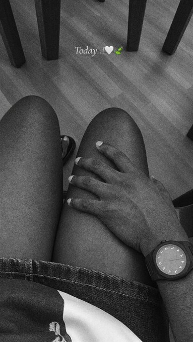 A black and white overhead photo showing a person's lap with their hand resting on their knee, wearing a wristwatch, with the text overlay 'Today... 🤍💚'.
