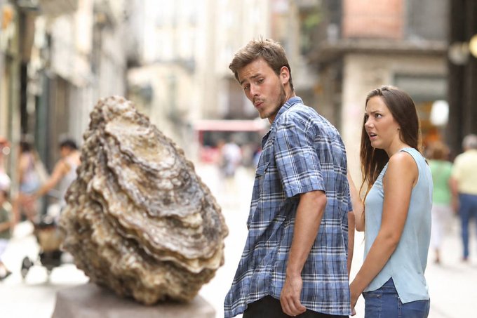 A man and a woman stand on a city street, both looking at a massive oyster shell prop with expressions of surprise or disgust.