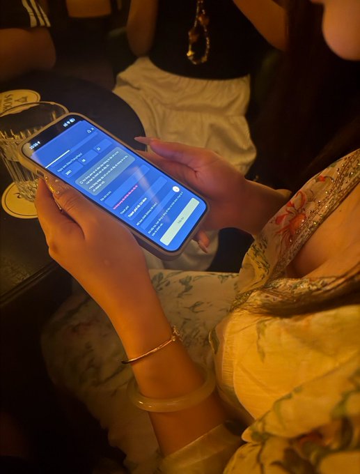 The image shows a woman holding up her phone, which has an app open on its screen. The app appears to be related to Binance trading or cryptocurrency, as indicated by the text "Ngồi hướng dẫn tạo tài khoản Binance của สมาย". The woman is wearing a floral dress and holding her phone in front of her face, which suggests that she might be engaged in an online conversation or browsing through the app.