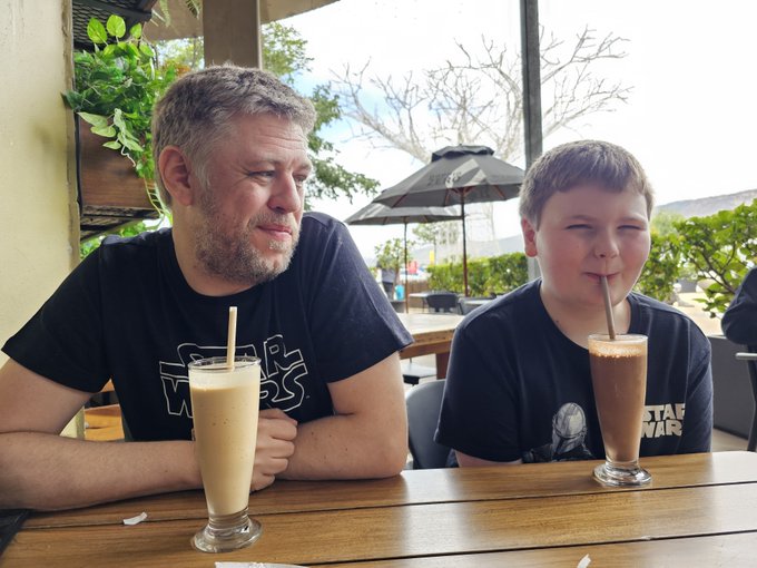 A man and a young boy wearing Star Wars t-shirts sit at a wooden table outdoors with two milkshake drinks.
