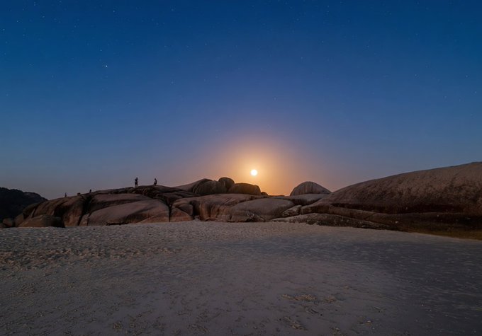 A nighttime photograph of a full moon rising over a rocky landscape with two small silhouettes of people standing on the boulders under a starry sky.