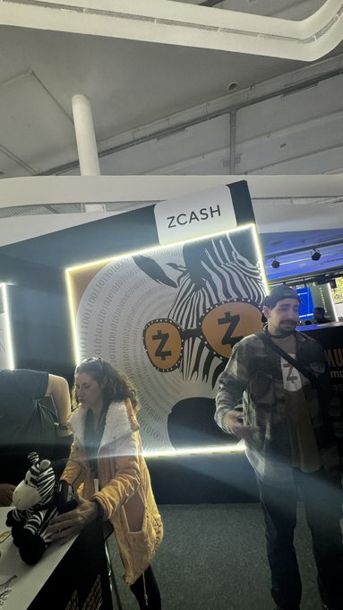 The image shows two people standing next to each other, with one person holding a stuffed animal while the other person has their hands behind their back. They are positioned near a booth or display area that features a zcash sign. The background of the image is dark and out of focus, drawing attention to the booth and its contents.