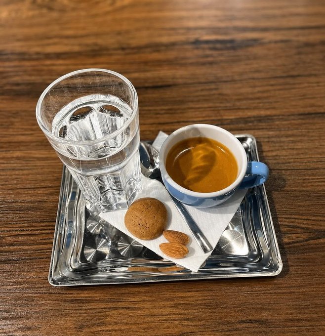 A small espresso cup with a blue rim, a glass of water, an almond cookie, and almonds sit on a silver tray atop a wooden table—a classic, energizing coffee break pairing that perfectly matches the post's "new week, new energy" vibe.