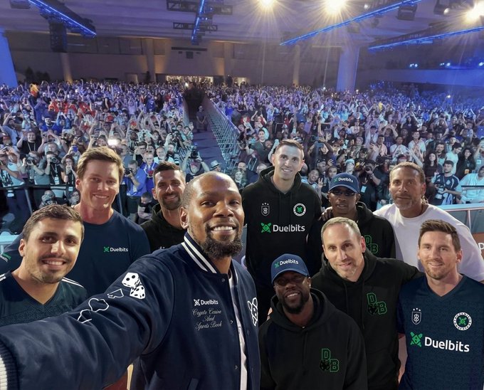 A group selfie of celebrities and soccer legends including Kevin Hart, Lionel Messi, and others wearing 'Duelbits' branded apparel, taken at a packed indoor arena event.