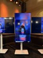 A photo of a display stand featuring a vertical poster or screen showing a face, placed in what appears to be an indoor venue.