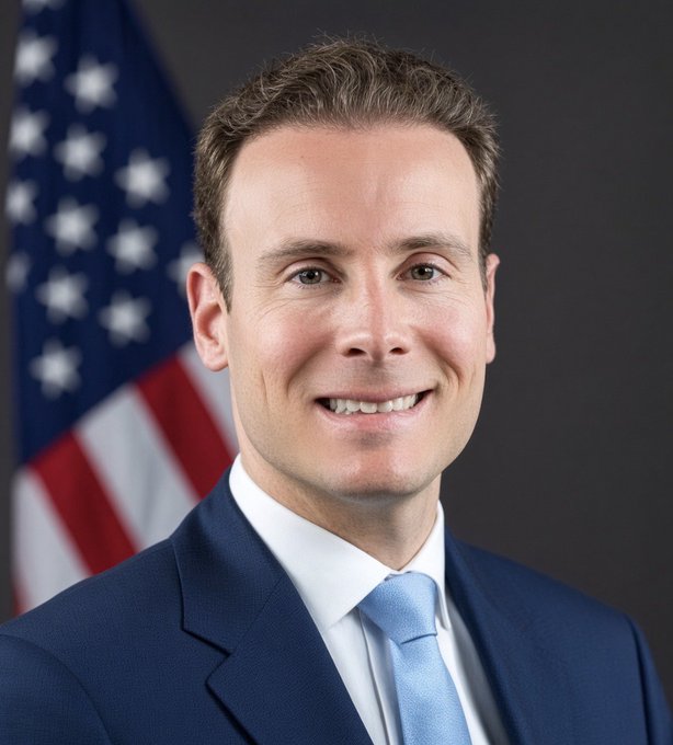 An official portrait of a smiling man in a blue suit and light blue tie standing in front of an American flag.