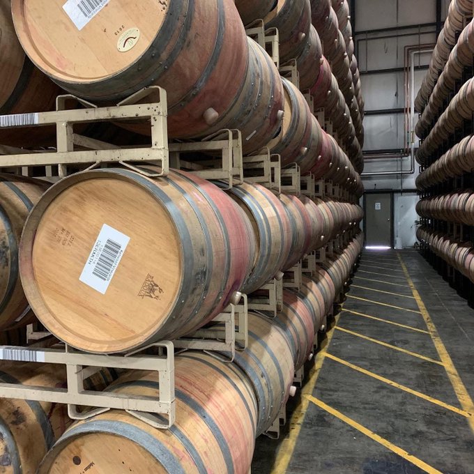 A wine cellar warehouse aisle lined with stacked wooden wine barrels on metal racks, each barrel bearing barcode labels, with yellow safety lines marking the walkway.