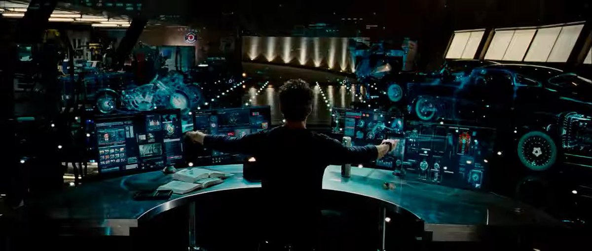 A person viewed from behind sits at a large console with multiple glowing screens displaying technical data and graphics in a futuristic control room.