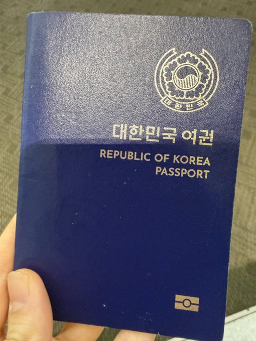 A person's hand holds up a dark blue Republic of Korea passport, featuring the Korean text "대한민국 여권" and the official emblem, suggesting they're traveling to Singapore for the TOKEN2049 conference.