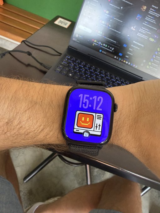A person wearing an Apple Watch with a blue retro TV smiley face watch face displaying the time 15:12, with a laptop open to Twitter/X in the background.