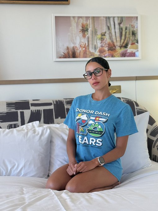 A woman wearing glasses and a blue "Donor Dash 50 Years: Hope, Legacy, Endurance" t-shirt sits on a white bed beneath a desert cactus artwork, visually tying into the post's motivational push to "get 1% better today."