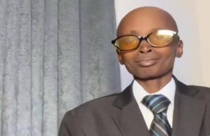 A bald man wearing yellow-tinted sunglasses, a dark suit, and a striped tie stands against a backdrop of gray curtains.