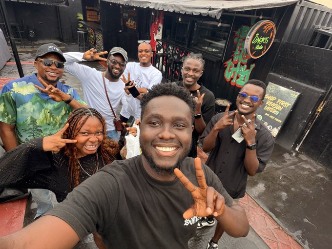 A group selfie of seven smiling people making peace signs outside what appears to be a bar or restaurant with neon signs including 'Cheers Bar' and 'DRINK FIRST REGRET LATER'.