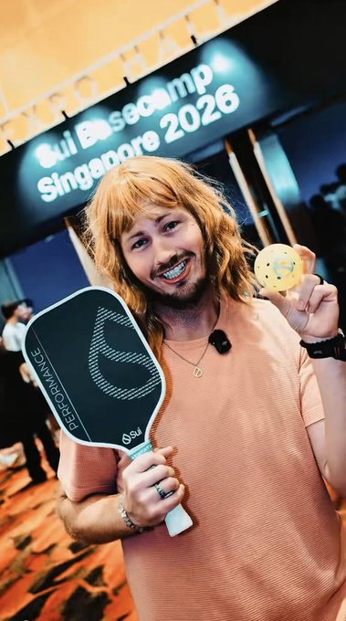 A person with long reddish-blonde hair smiles while holding a pickleball paddle and a perforated ball, standing in front of a banner reading 'Gui Basecamp Singapore 2026'.