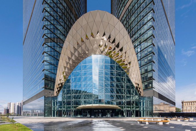 A modern glass and steel building with a distinctive curved archway over its entrance, reflecting the blue sky and surrounding cityscape.