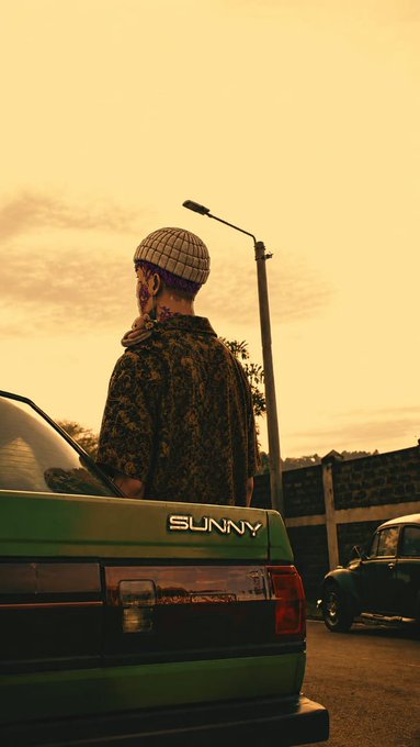 Cinematic photo of a person with facial tattoos, beanie, and patterned coat standing by a green car labeled "SUNNY" at sunset, with a vintage Volkswagen Beetle in the background.