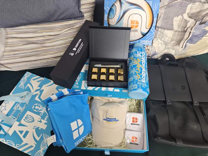 An opened gift box reveals a World Cup-themed merchandise package from DeepCoin (深币), featuring a blue and white Argentina-themed backpack, jersey, custom ice packs reading "DEEP COIN," and an elegant set of golden pins, matching the poster's description of receiving a co-branded World Cup gift haul.