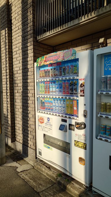 A photograph of a Japanese beverage vending machine situated outside against a tan brick wall in a narrow alleyway.