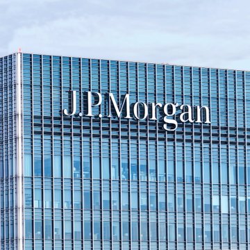 A modern glass skyscraper prominently displays the "J.P.Morgan" logo across its facade, representing the financial giant weighing in on Bitcoin's potential price support relative to gold. The image visually anchors the bank's analysis on cryptocurrency versus gold ETF dynamics, as suggested by the headline commentary.