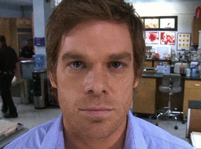 A man with short brown hair wearing a light purple shirt stares directly at the camera in what appears to be a lab or office setting, with monitors visible in the background.