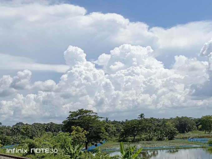 A landscape photo taken with an Infinix Note 30 showing dramatic white clouds over lush green trees and a small body of water.