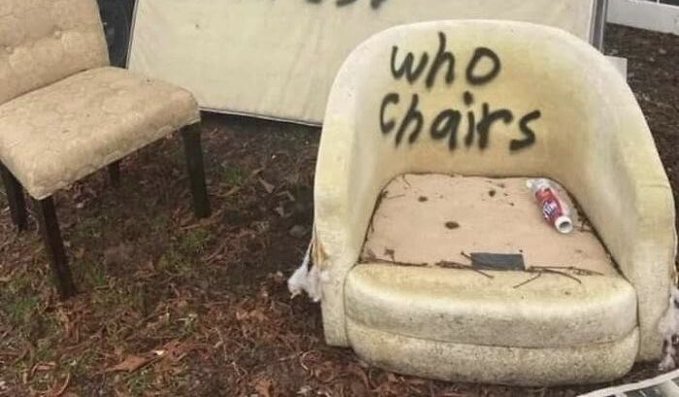 A discarded, dirty cream-colored car seat sitting on grass with "who chairs" spray-painted on the headrest and a spray paint can resting on the seat cushion.