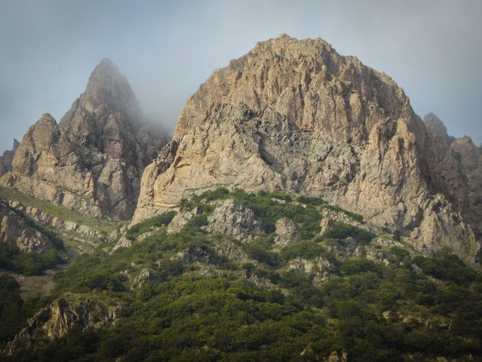 A rugged, mist-kissed mountain rises above lush green vegetation under a hazy sky, capturing the dramatic natural scenery of the Chalus Road region. The image embodies the ethereal, untamed beauty celebrated in the "Mystical Peaks of Chalus Road" NFT collection.